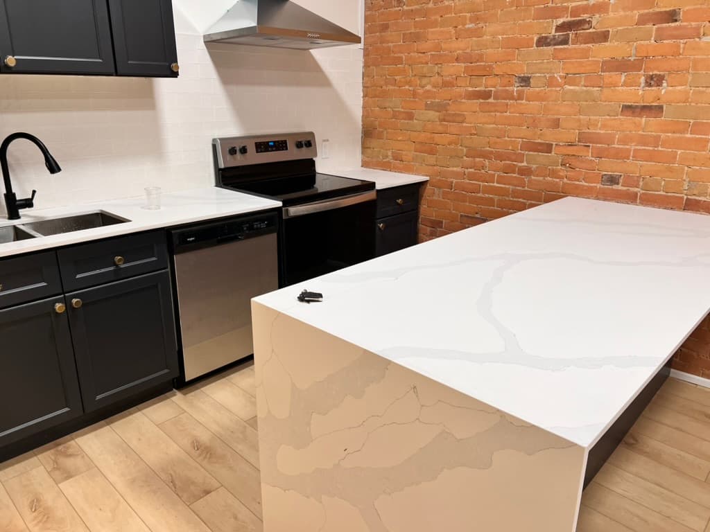 A kitchen with dark cabinets, a white quartz waterfall island and an exposed brick wall
