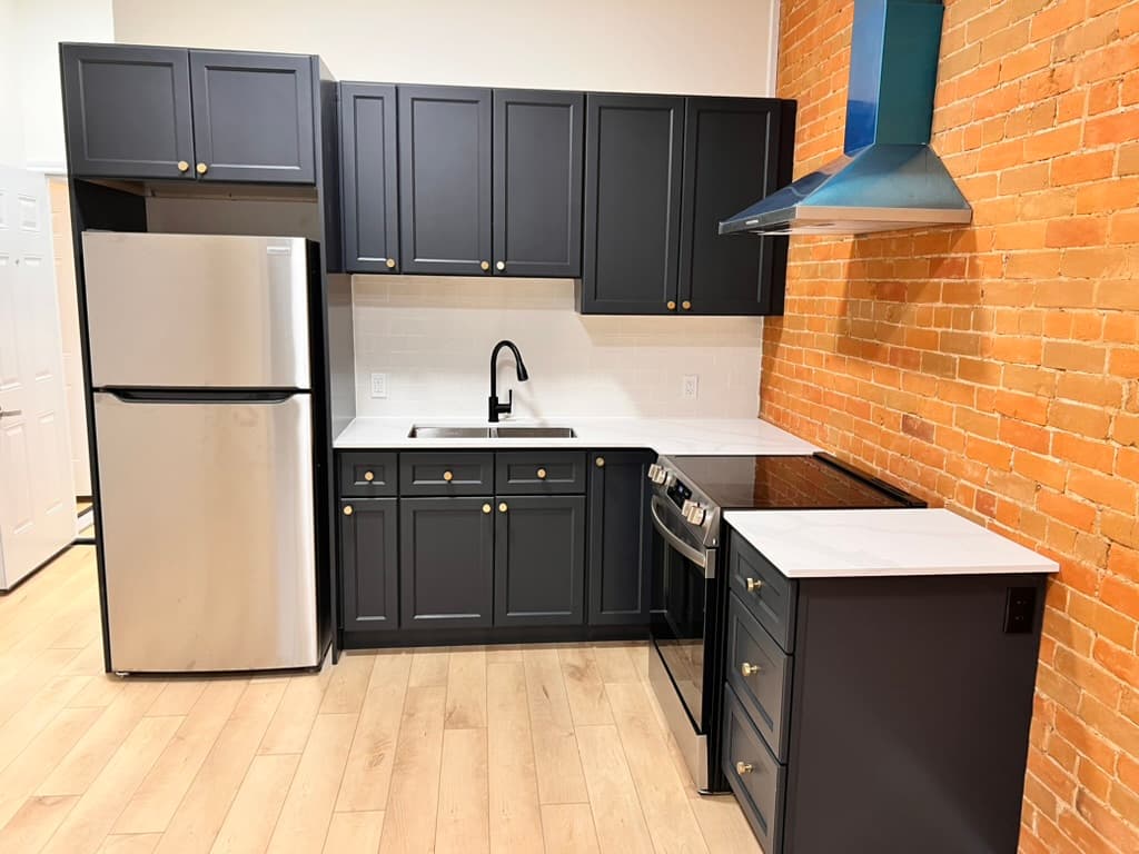 A compact kitchen with dark cabinets, brass handles and a stainless steel fridge beside an exposed brick wall
