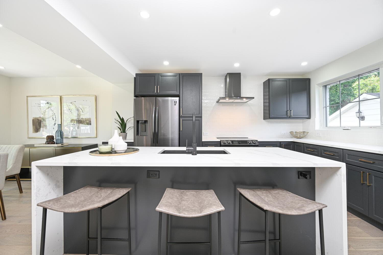 A white waterfall island with three stools in front of dark cabinets and stainless appliances