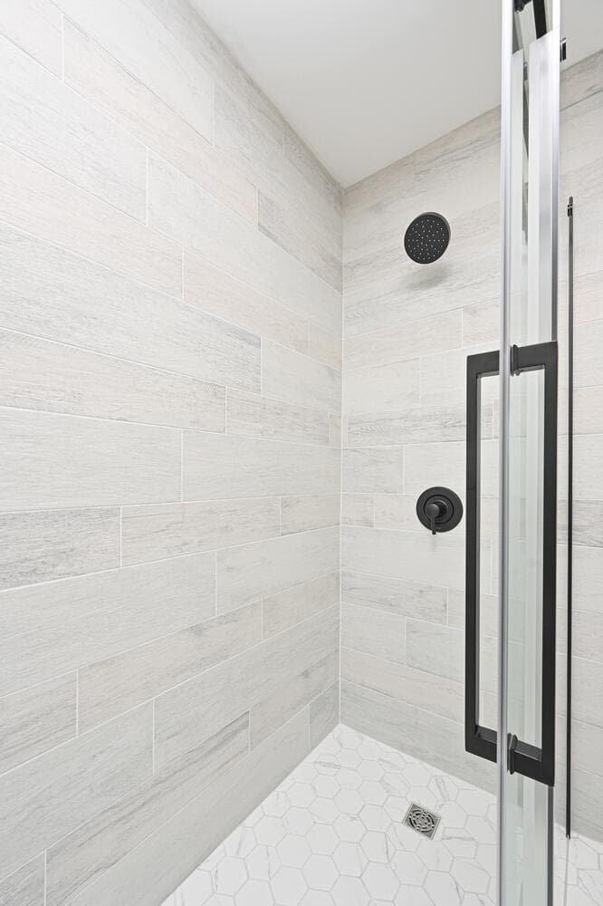 A walk-in shower with a sliding glass door, wood-look wall tile and a white hexagon floor