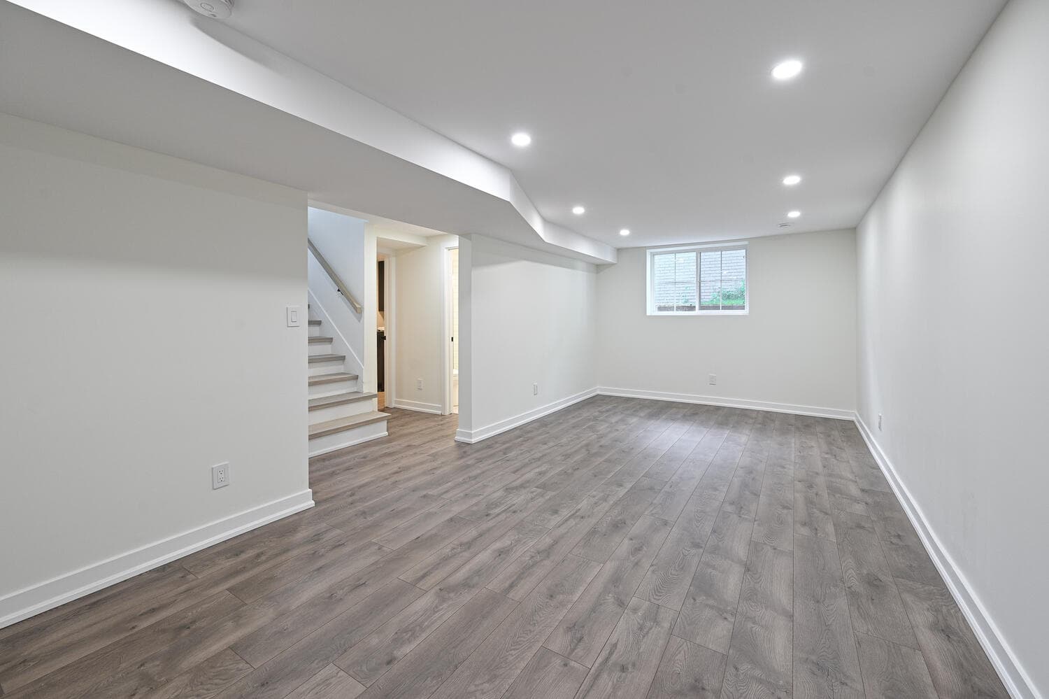 A finished basement room with grey wood-look floors, white walls, pot lights, a small window and stairs leading up