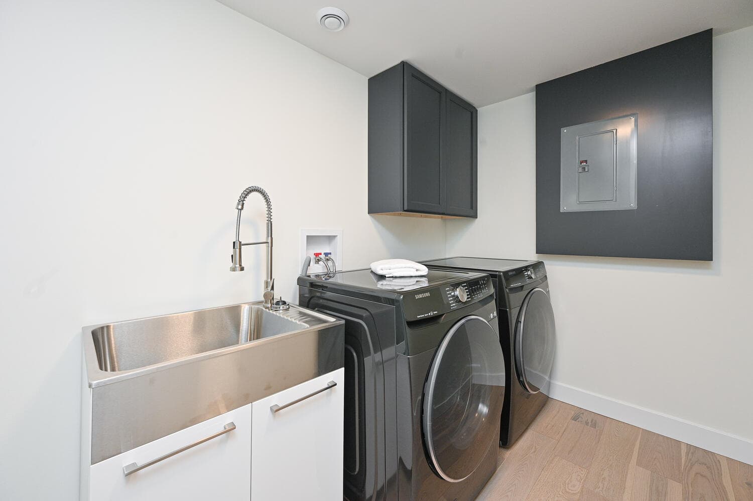 A laundry room with a front-load washer and dryer, a deep steel sink and dark upper cabinets