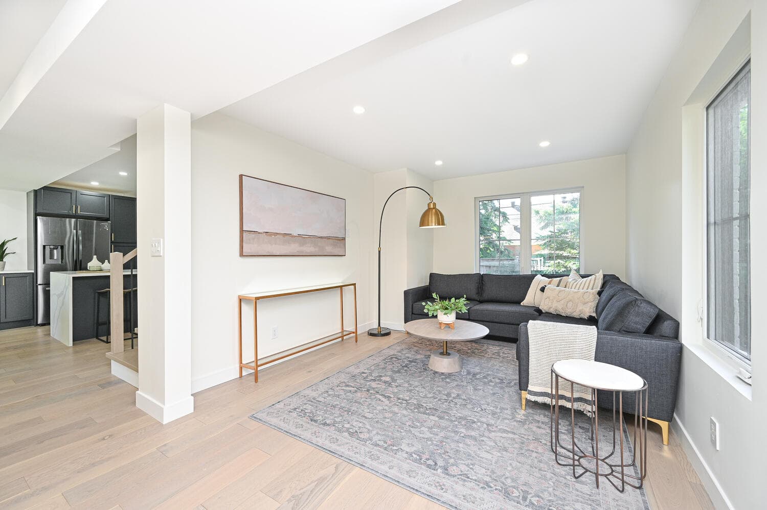 An open living room with a grey sectional sofa and light oak floors, with the kitchen visible beyond