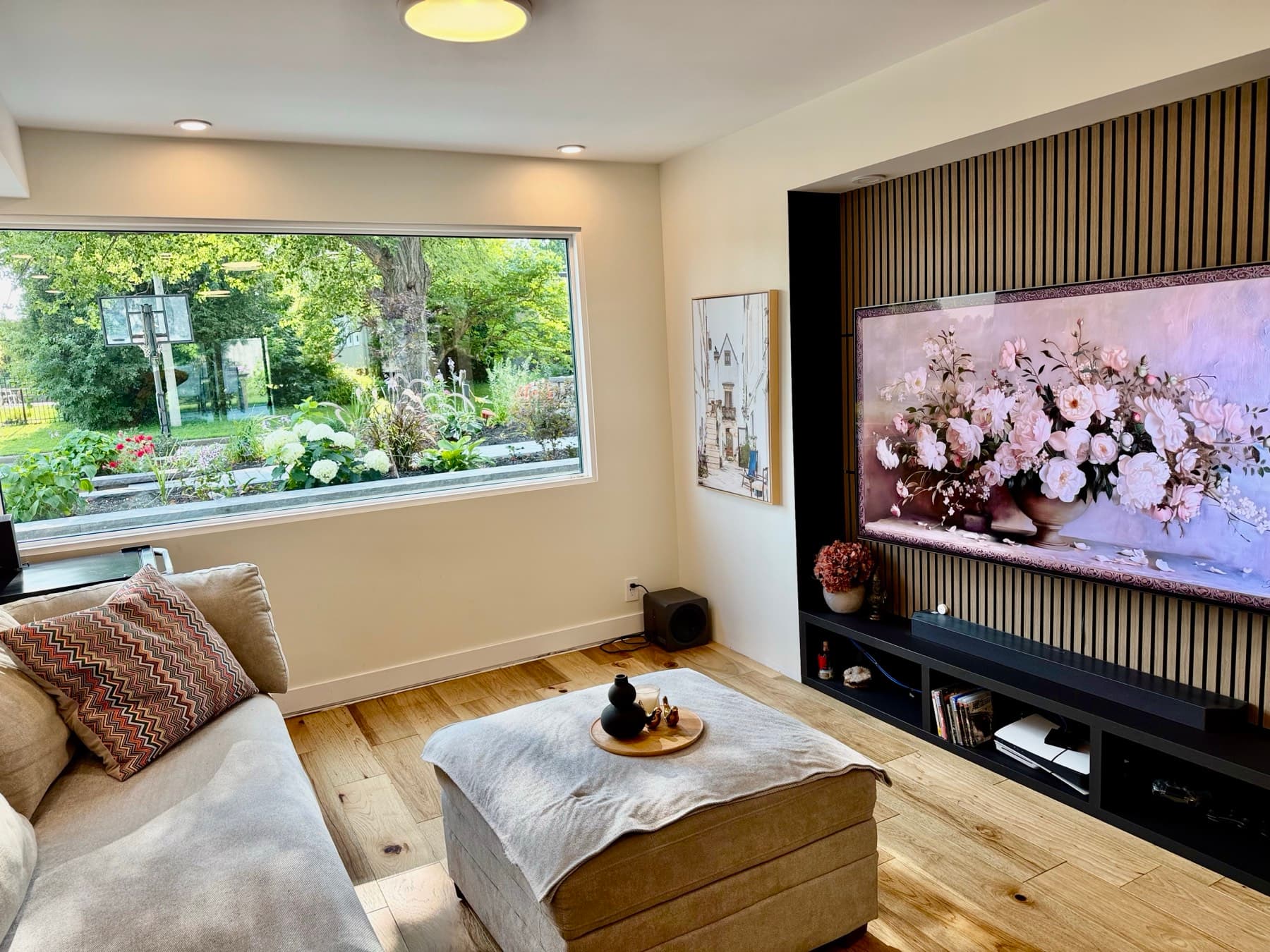 A family room with a sofa and ottoman, a slatted wood media wall and a wide window onto a flowering garden