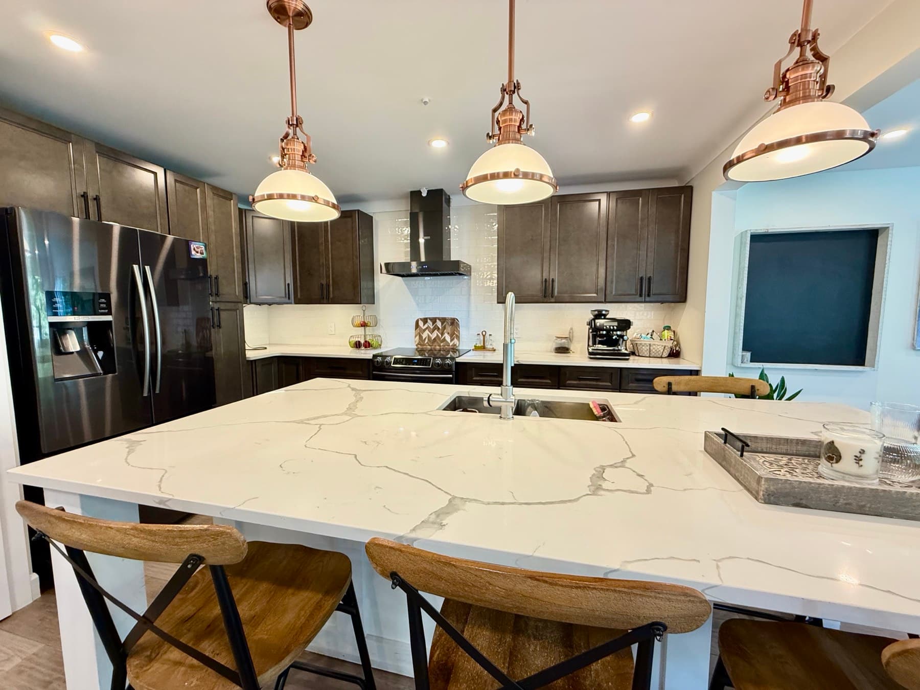 A kitchen with a white quartz island, three copper pendant lights, dark cabinets and a white tile backsplash