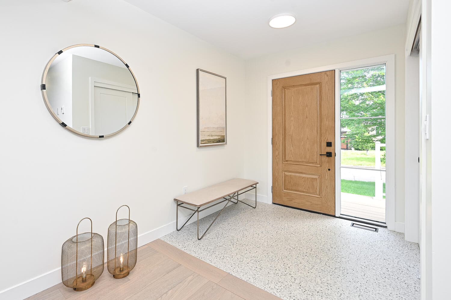 A bright front entry with a wood front door and a glass side panel, a bench and a round mirror