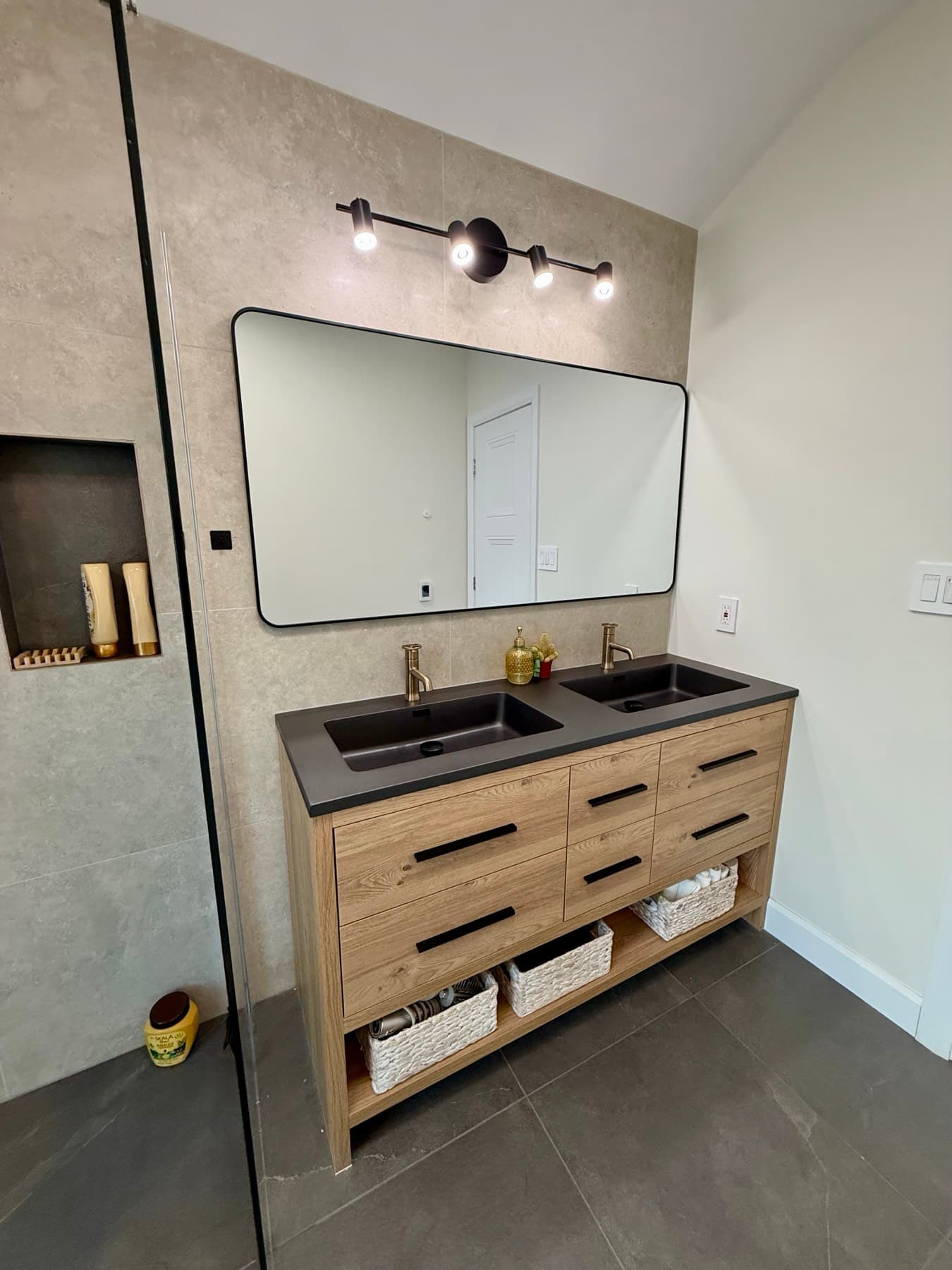 A wood double vanity with two dark basins under a wide mirror, beside a tiled shower with a wall niche