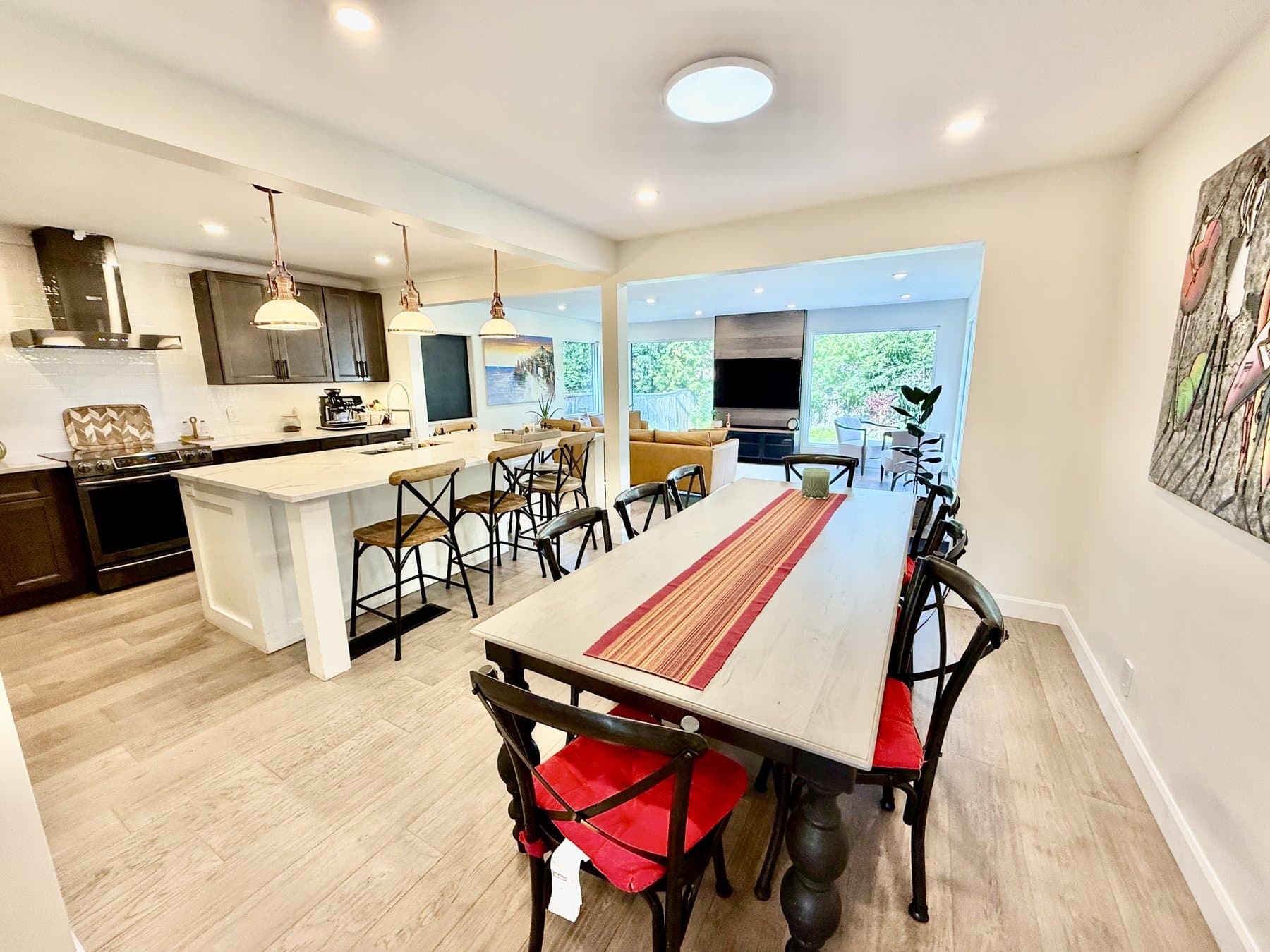 A long dining table with red cushioned chairs beside the kitchen island, with the living room and its large windows beyond