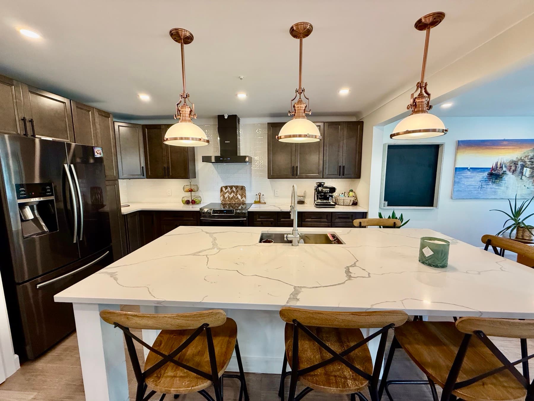 A kitchen with a large white quartz island, copper pendant lights and dark cabinets